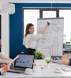 Company head standing in front of audience diverse staff members making presentation. Serious speaker boss executive, business trainer explaining development strategy to motivated mixed race employees.